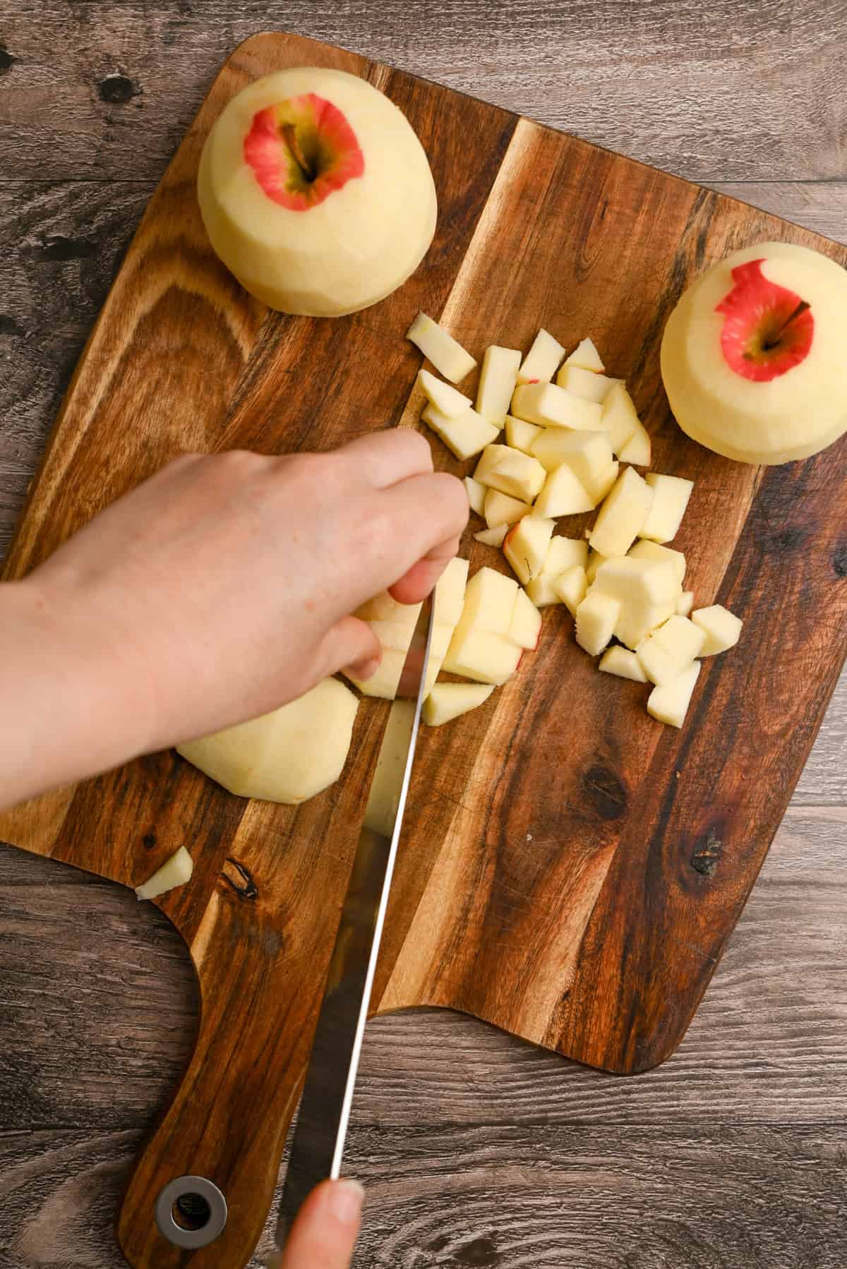 A hand is dicing a peeled apple with a knife on a wooden cutting board, while two other peeled apples with some skin remaining on top sit nearby.