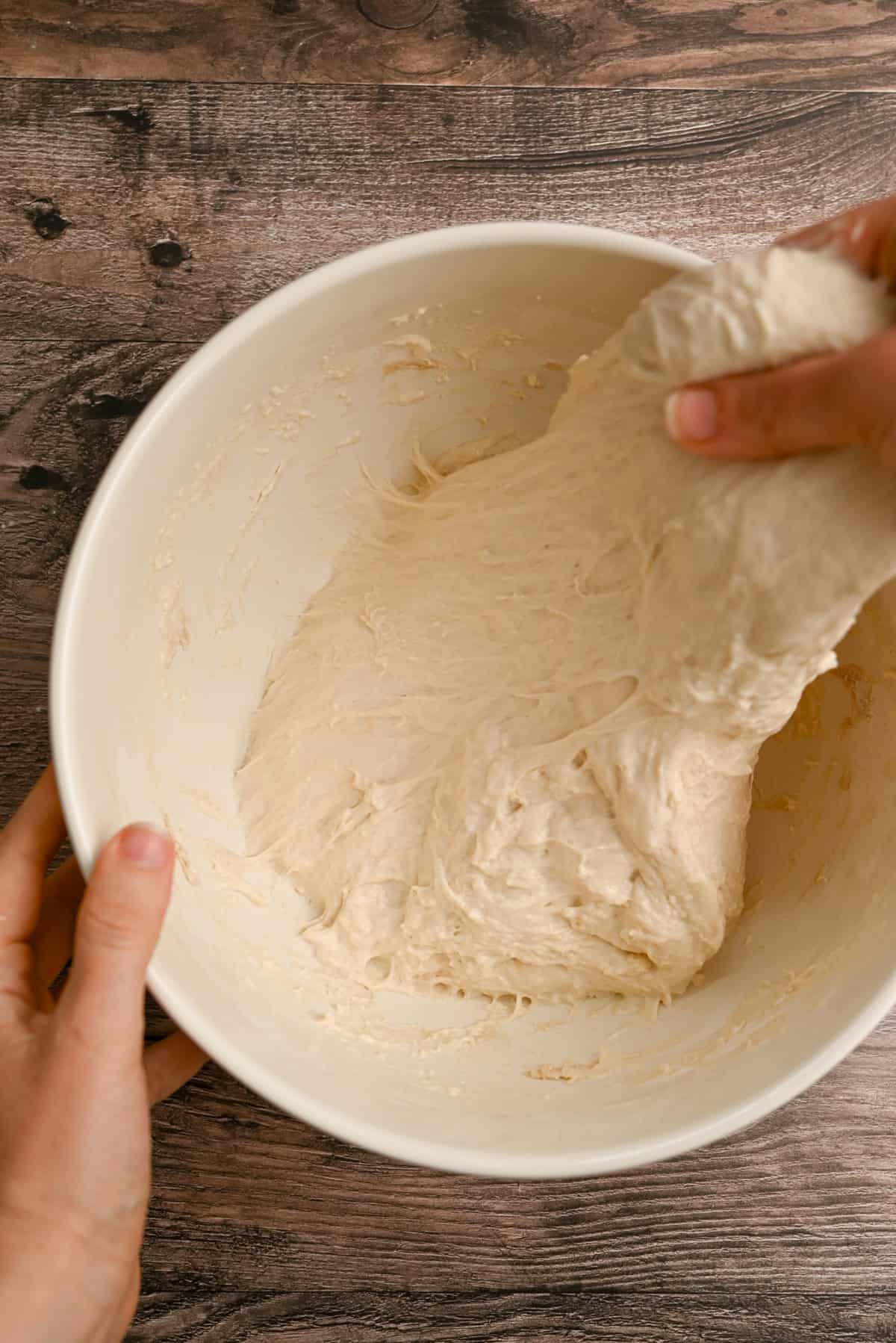 A person stretches sticky bread dough in a large white mixing bowl on a wooden surface.