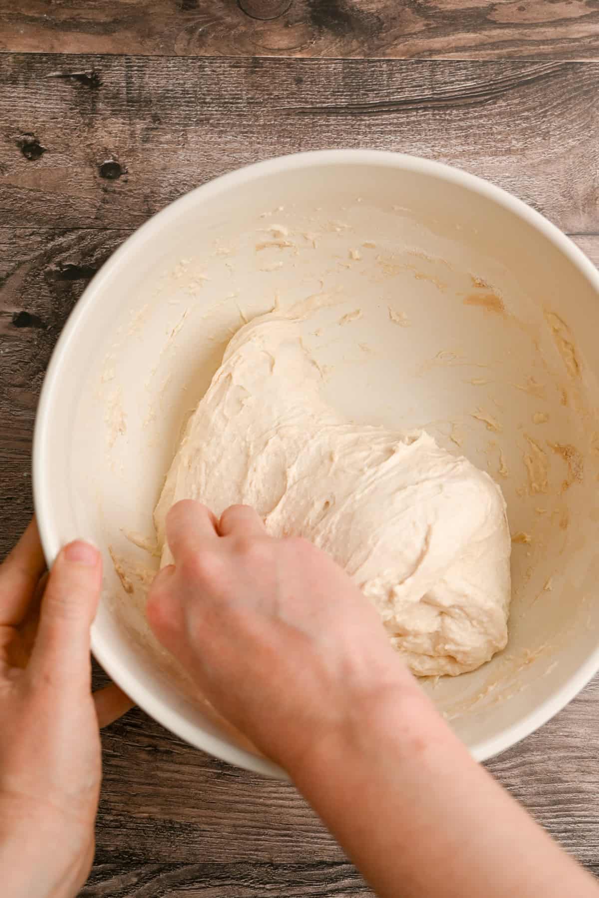 A person kneads soft dough in a large white mixing bowl on a wooden surface.