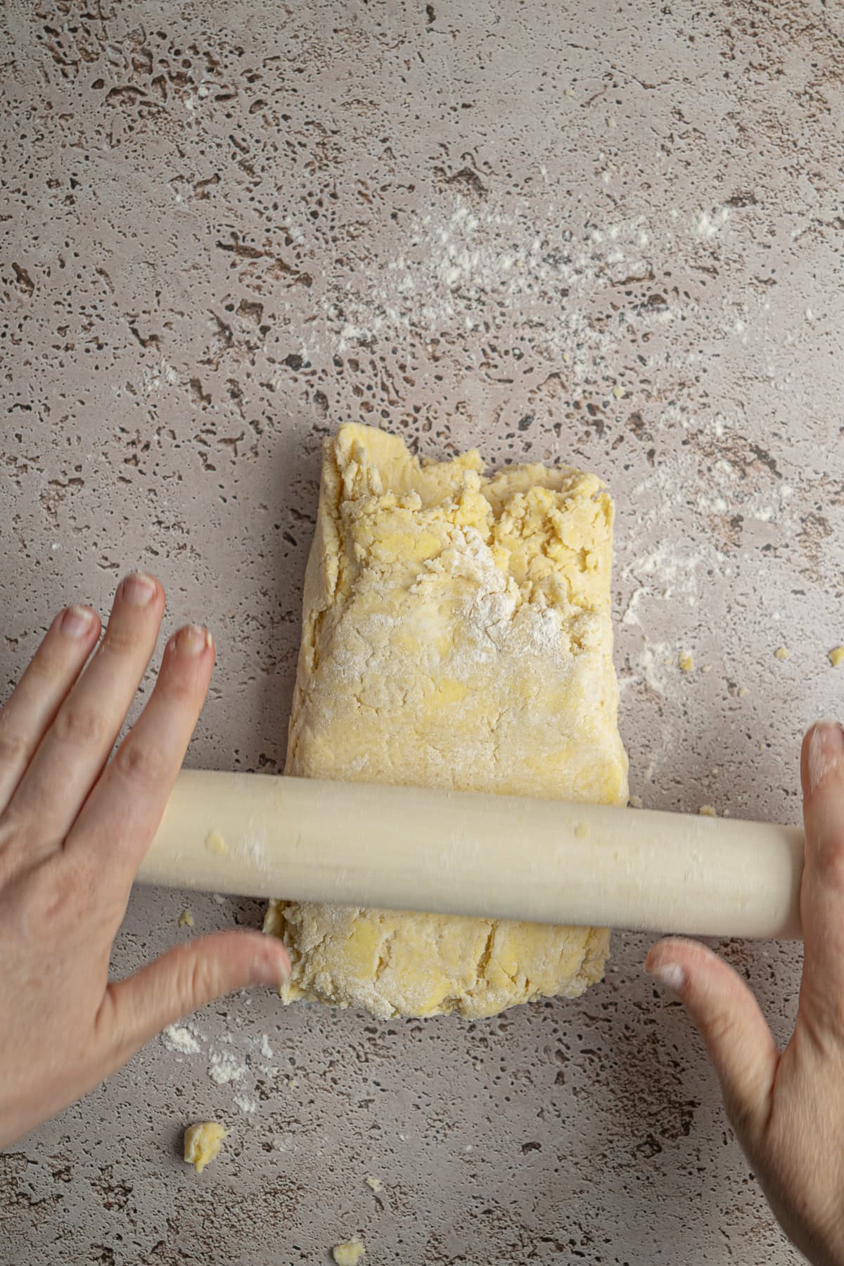 Hands use a rolling pin to flatten dough for Blackberry Hand Pies on a lightly floured, speckled countertop.