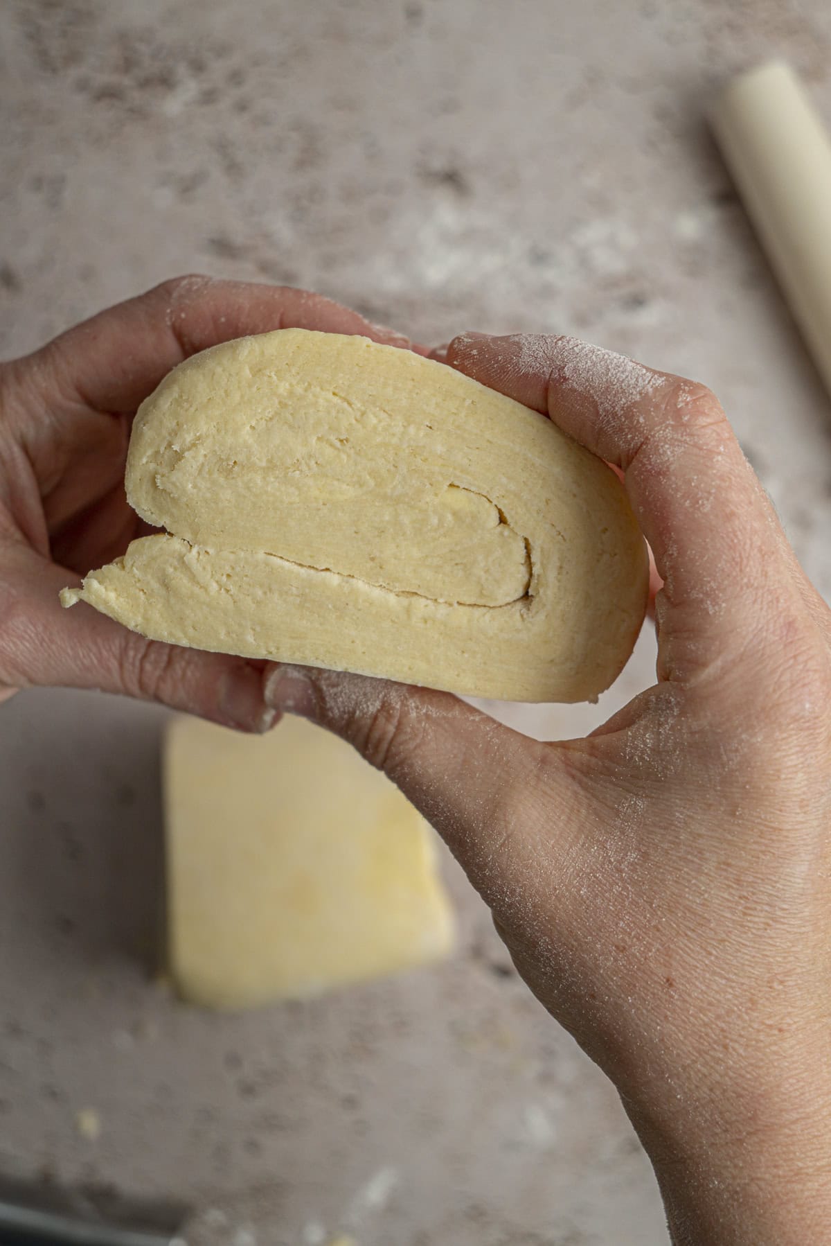 A close-up of two hands holding a rolled piece of dough, showing its layers. The surface around them is dusted with flour, capturing the beginning stages of making Blackberry Hand Pies.