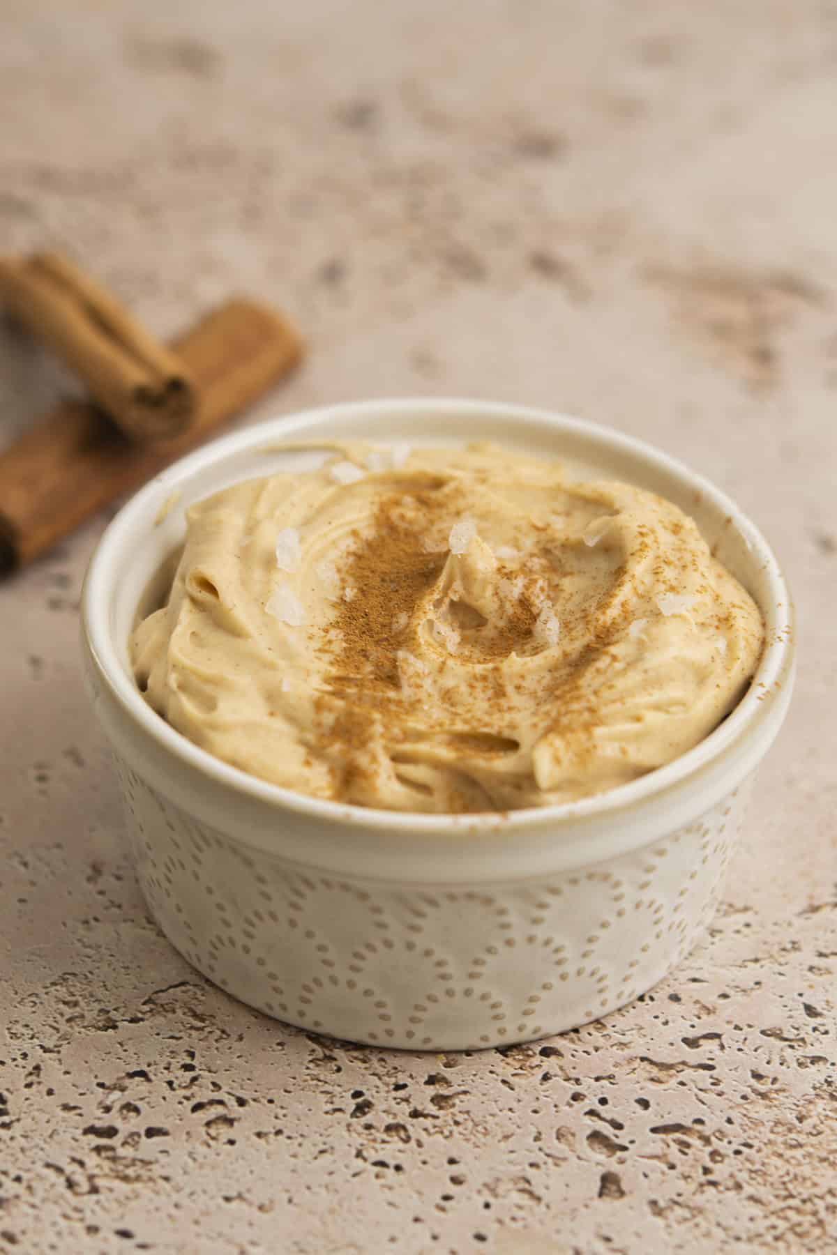A small white ramekin filled with creamy light brown dip, topped with a sprinkle of cinnamon, sits on a textured surface with two cinnamon sticks in the background.