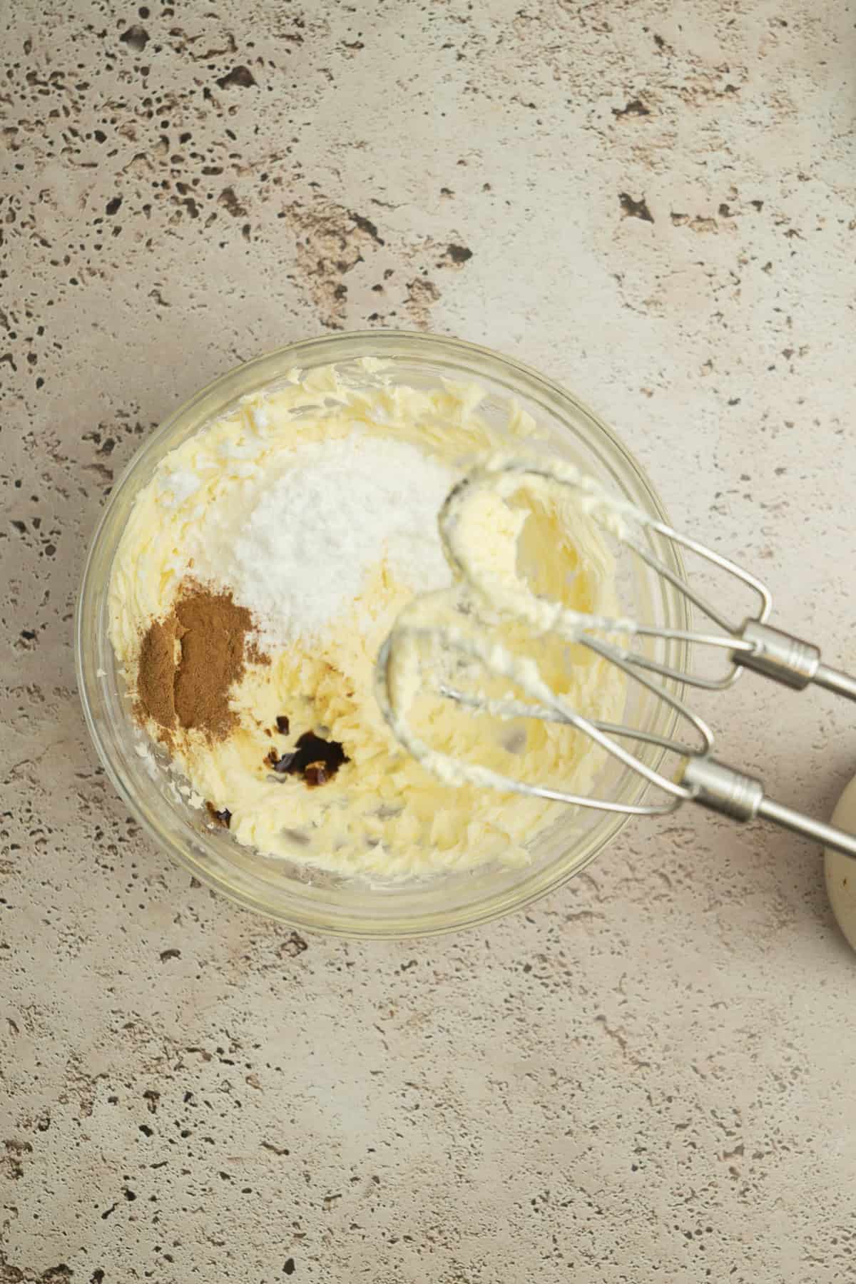 A glass bowl with creamed butter, sugar, cinnamon, and vanilla, next to a hand mixer with beaters, sits on a speckled beige countertop, ready for baking.