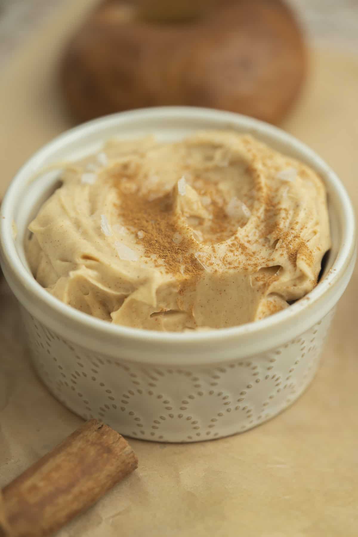 A small white ramekin filled with creamy whipped spread, sprinkled with cinnamon, sits on parchment paper. A cinnamon stick and a blurred baked good appear in the background.