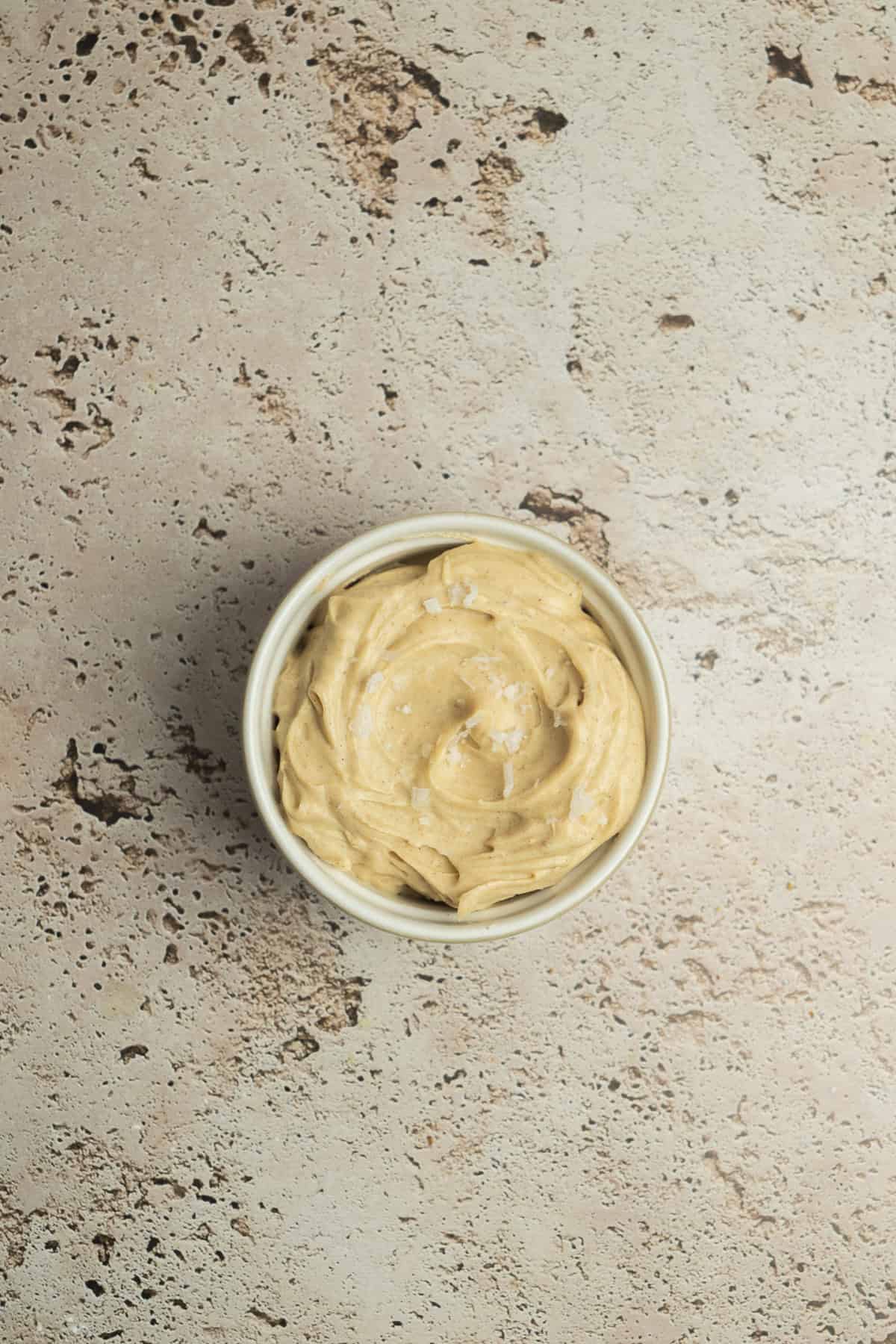 A small white bowl filled with creamy, light brown peanut butter on a textured beige surface.