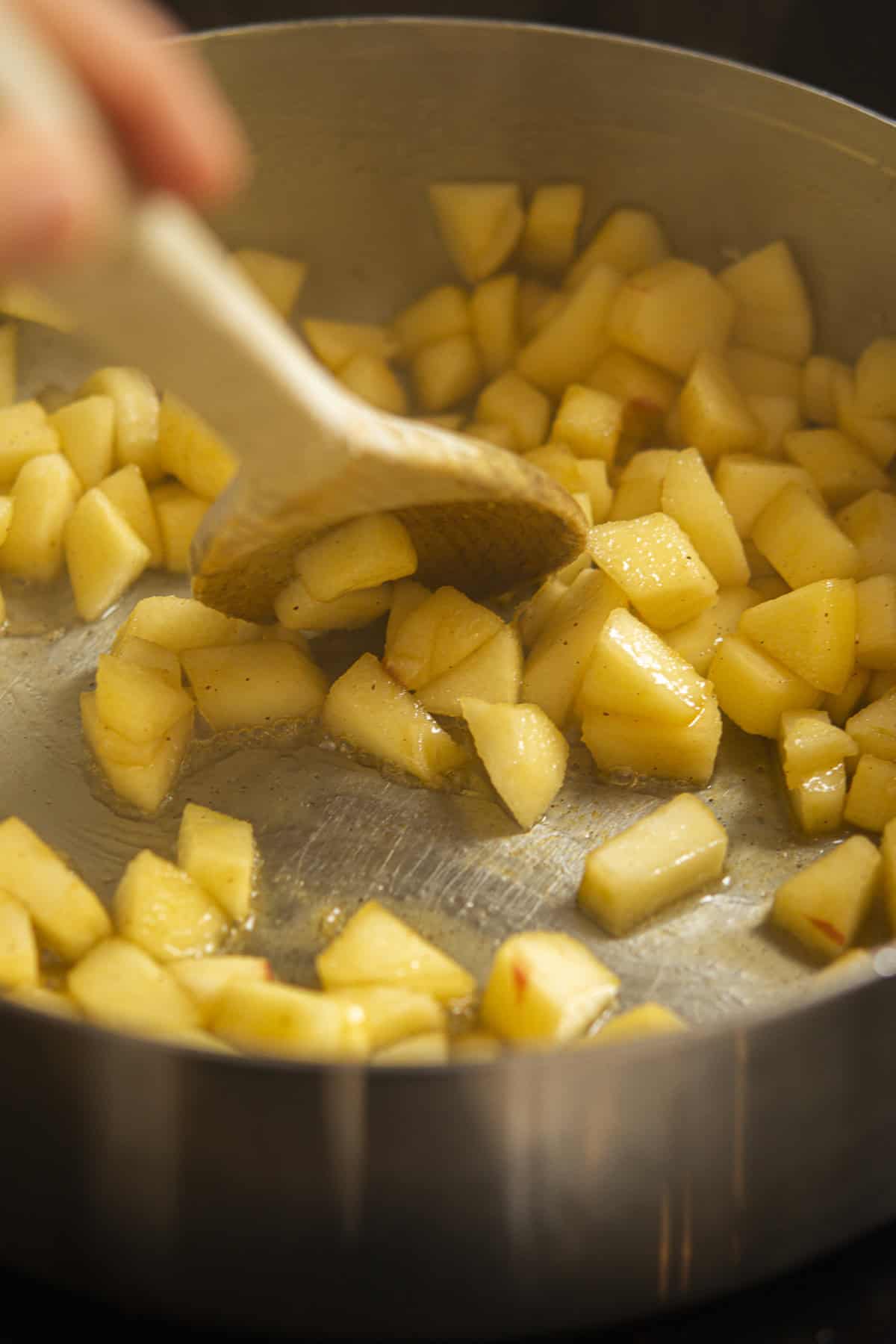 Diced apples being stirred with a wooden spoon in a pan, cooking and coated in a glossy mixture—perfect for topping warm pumpkin focaccia.
