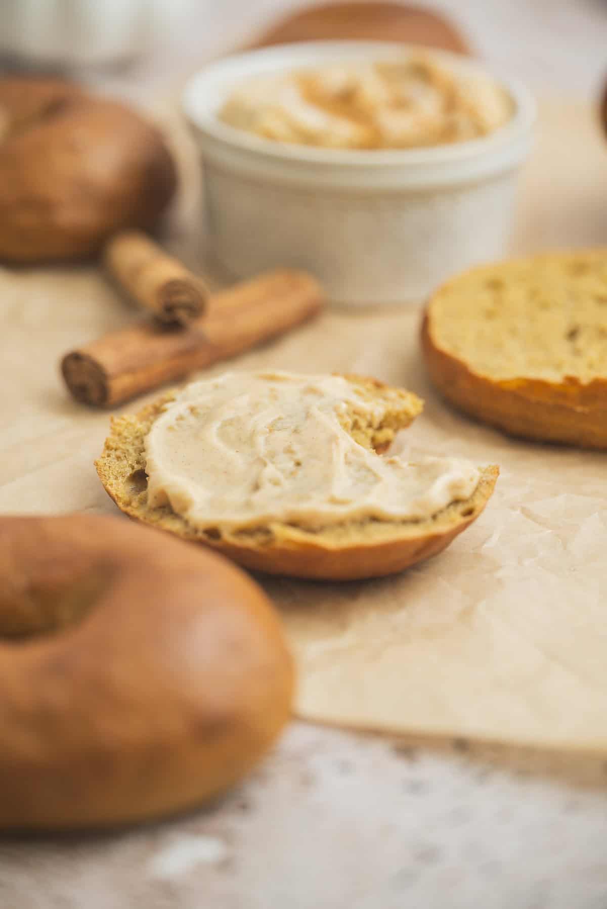 A sliced bagel with cinnamon cream cheese spread on one half sits on parchment paper, surrounded by whole bagels, cinnamon sticks, and a small bowl of spread in the background.