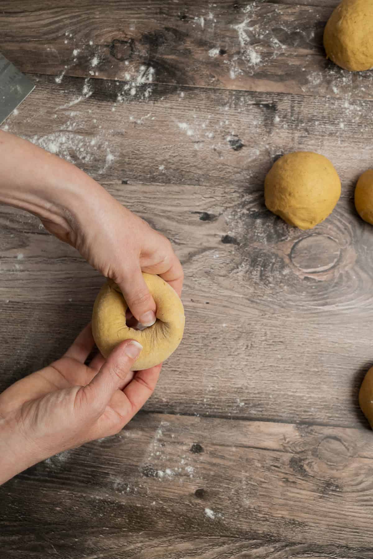 A person shapes a ball of dough by pressing a thumb through its center on a floured wooden surface, with several other dough balls nearby.