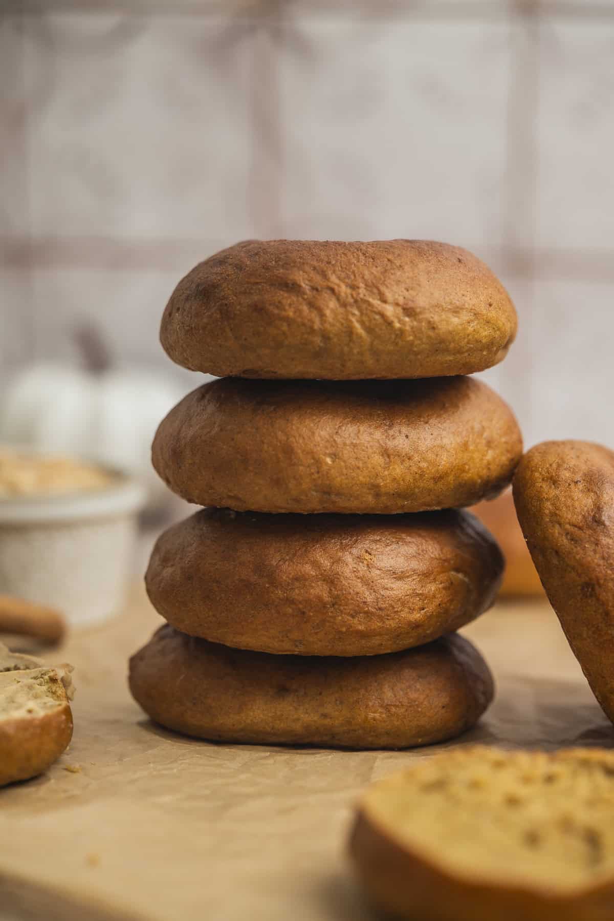 A stack of four golden-brown bagels sits on parchment paper, with a partial bagel and a bowl in the background, against a blurred tiled wall.