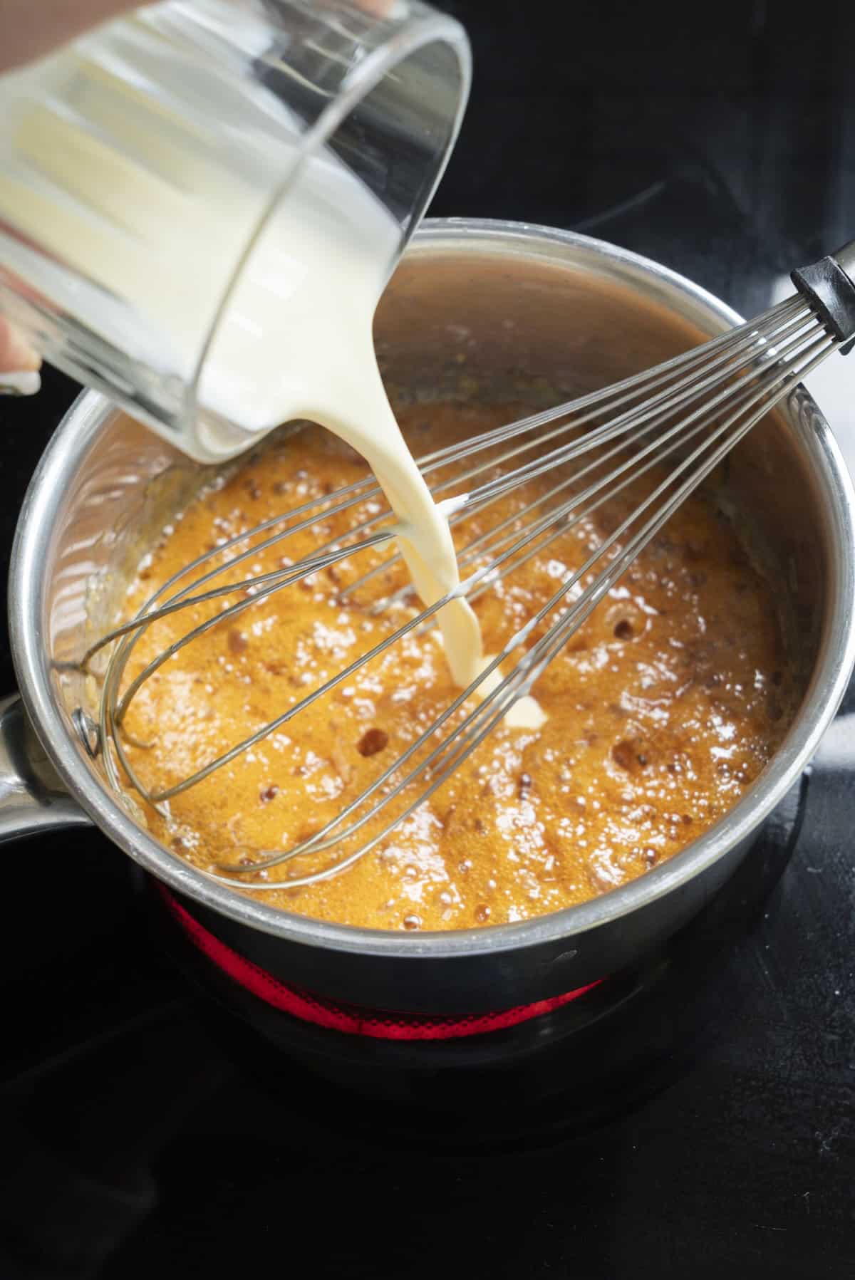A glass measuring cup pours cream into a saucepan of caramelized sugar for Cinnamon Caramel Ice Cream, with a metal whisk resting inside, on a stovetop.