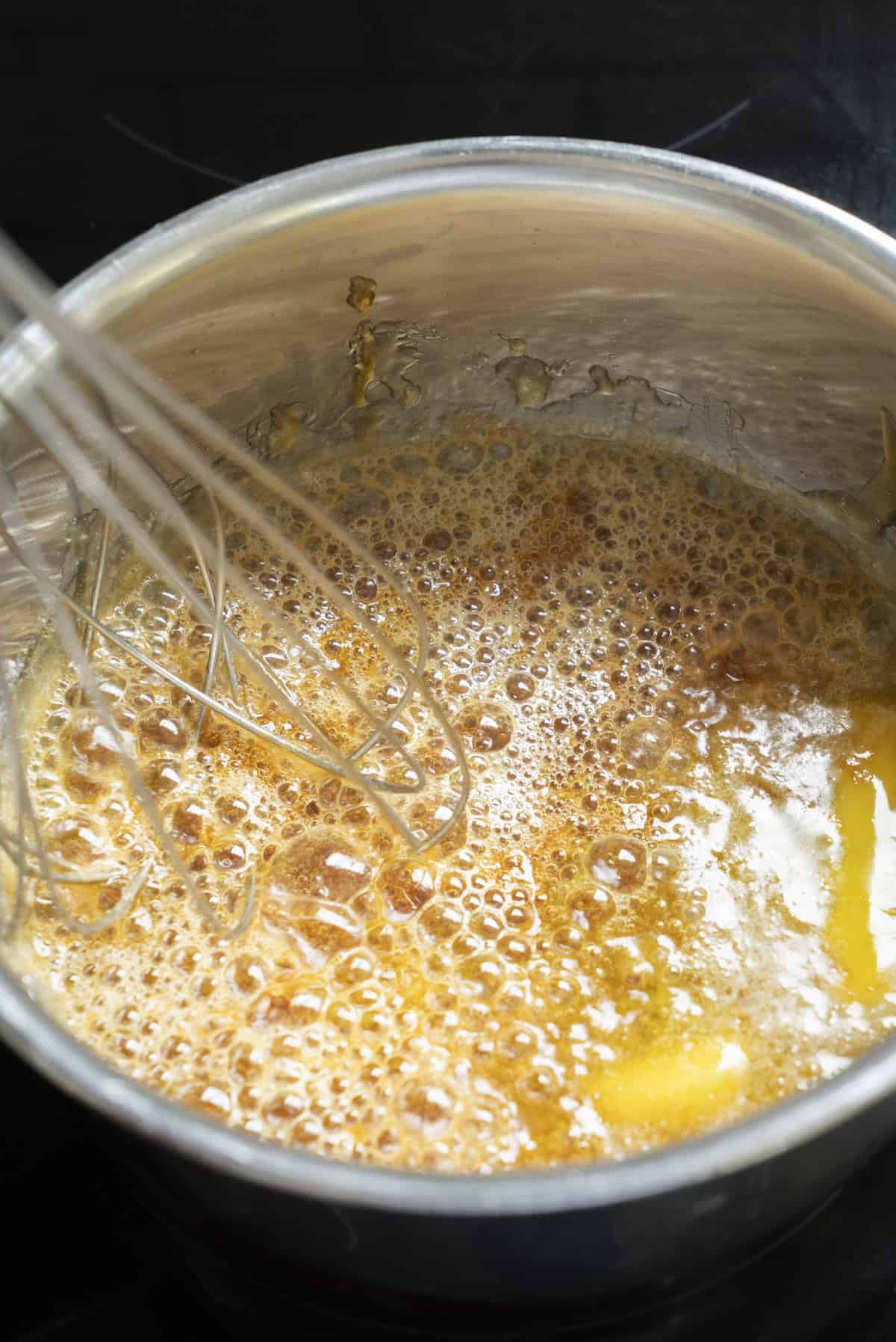 A metal whisk stirs bubbling, golden-brown caramel sauce for Cinnamon Caramel Ice Cream in a stainless steel pot on a stovetop.