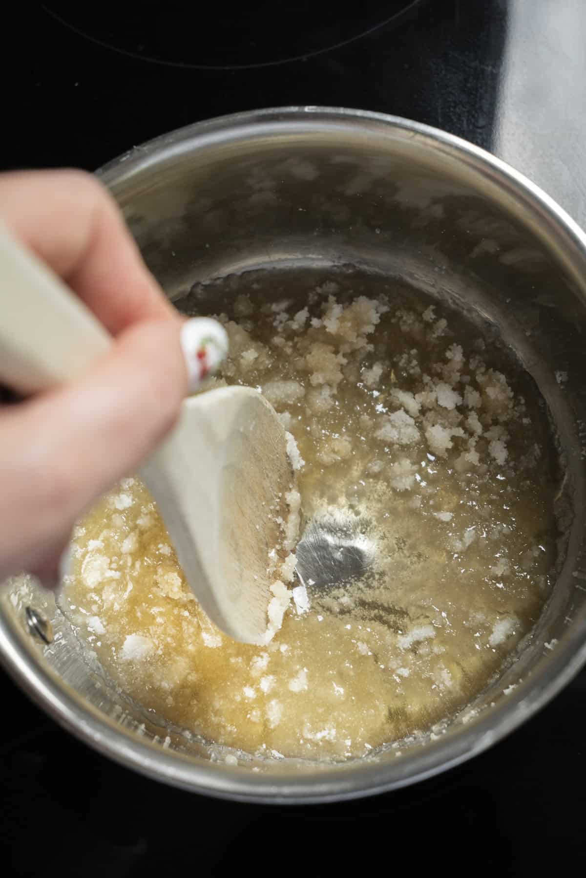 A hand stirs a mixture of sugar and melted butter in a stainless steel saucepan with a wooden spoon, as the base for Cinnamon Caramel Ice Cream begins to dissolve and blend together.