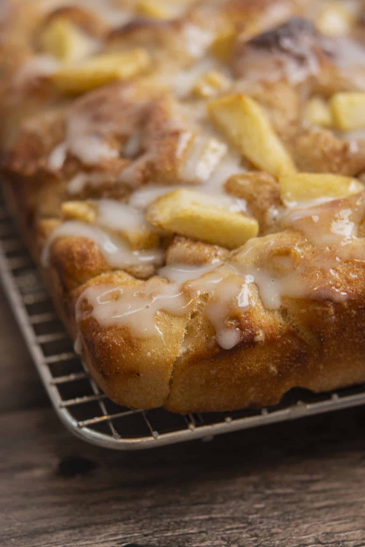 Close-up of a glazed apple cake with chunks of apple on top, resting on a wire cooling rack over a wooden surface.