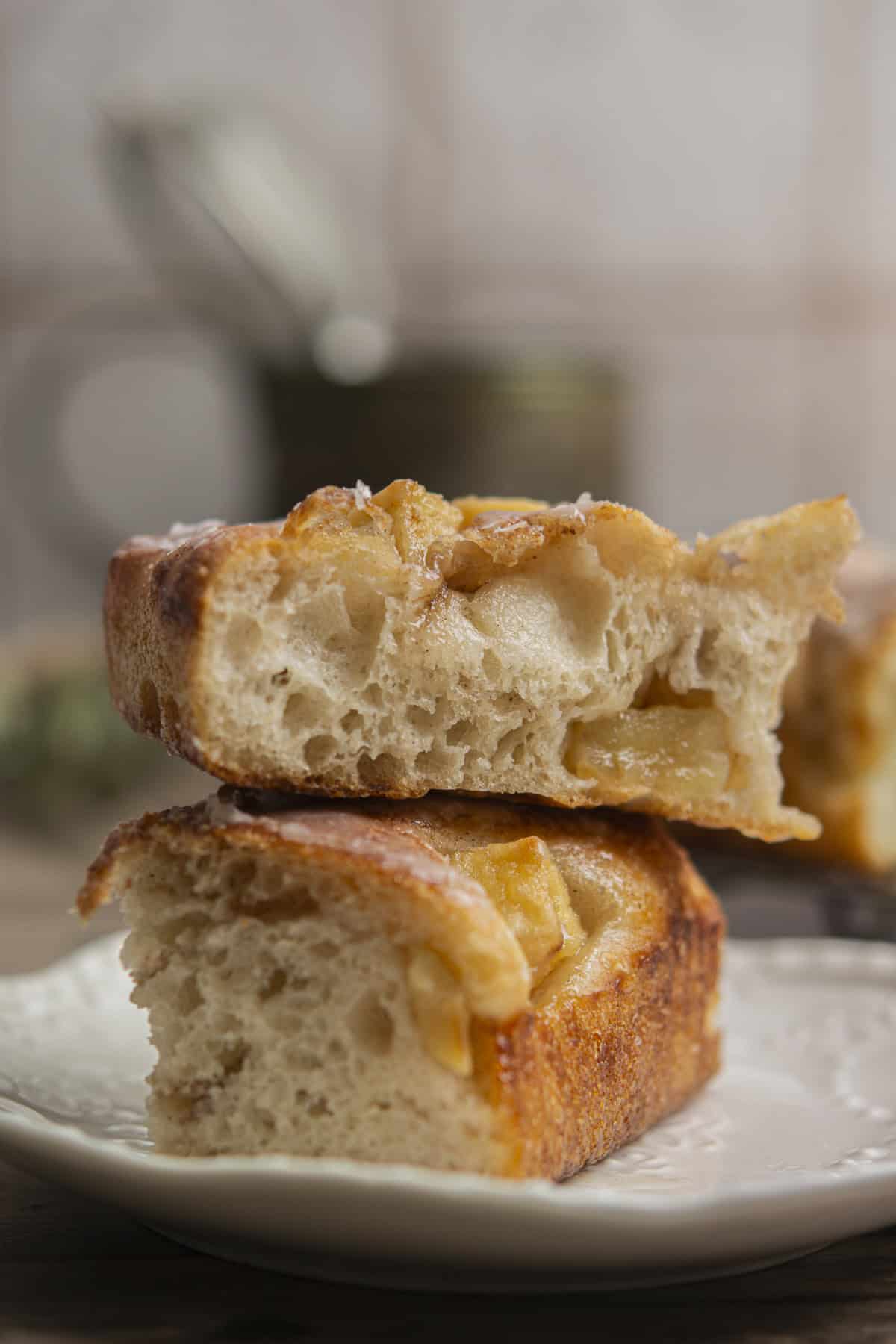 Two pieces of thick, golden-brown focaccia bread with visible apple chunks are stacked on a white plate, showing their soft, airy texture and caramelized top. The background is softly blurred.