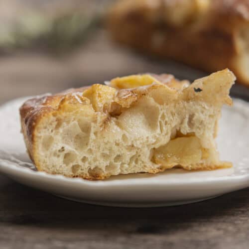 A close-up of a slice of airy, golden-brown bread with visible chunks of baked apples on a white plate, sitting on a wooden surface. Another slice is blurred in the background.