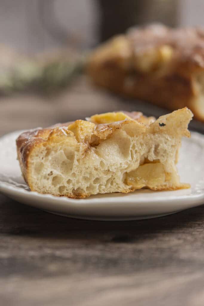 A close-up of a slice of airy, golden-brown bread with visible chunks of baked apples on a white plate, sitting on a wooden surface. Another slice is blurred in the background.