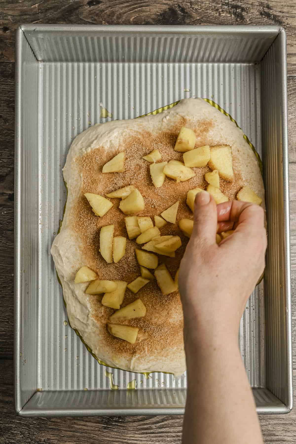 A hand sprinkles chopped apples onto dough covered with cinnamon and sugar in a rectangular metal baking pan on a wooden surface.