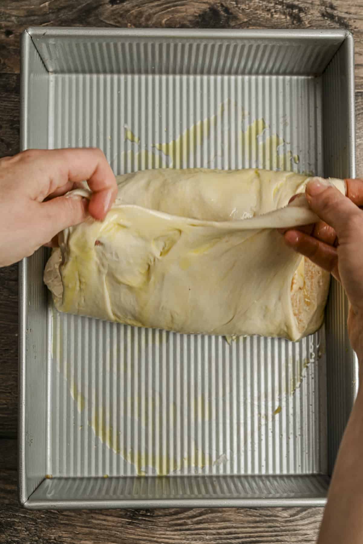 Hands folding dough over a loaf in a greased rectangular baking pan on a wooden surface, preparing it for baking.