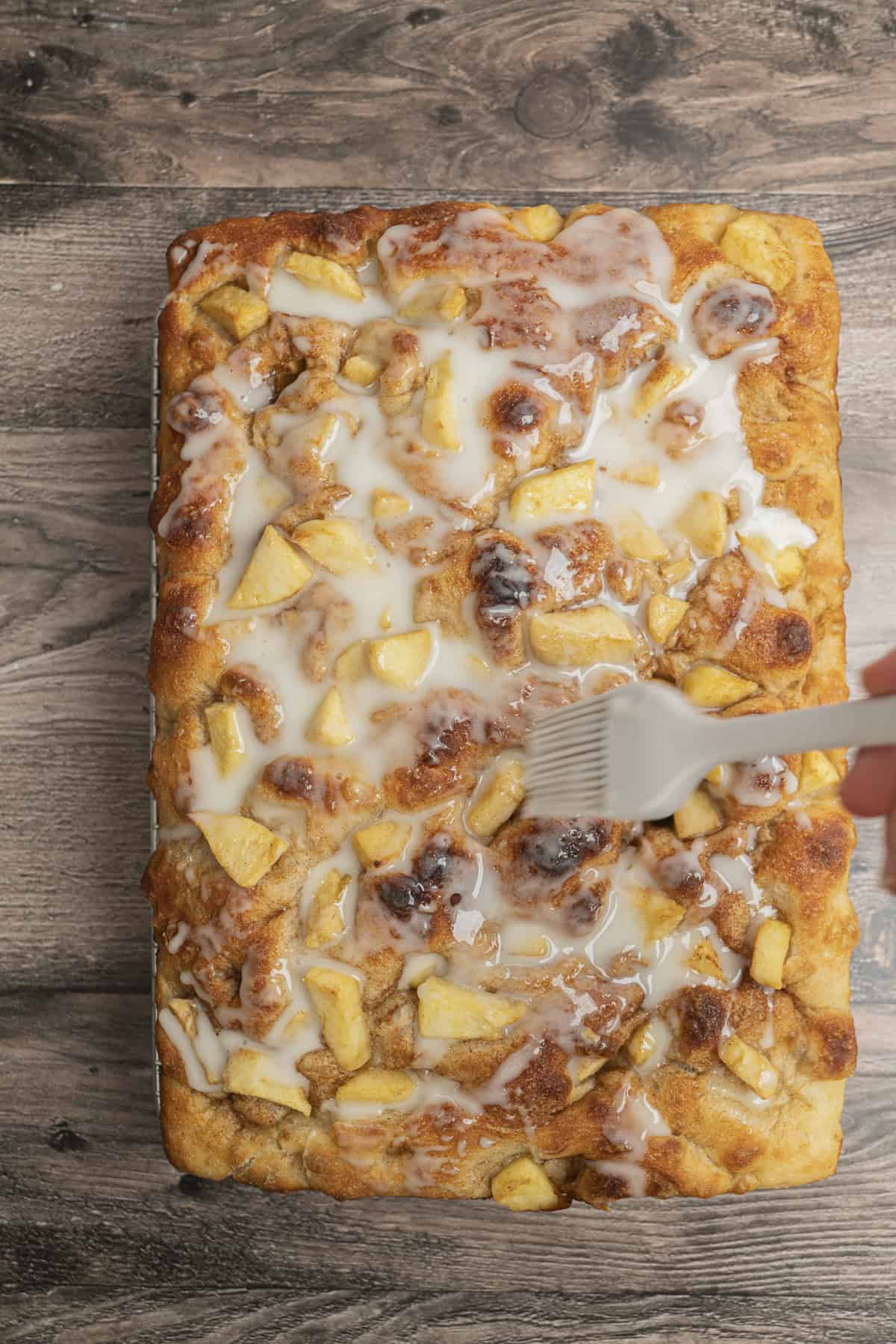 A hand uses a brush to spread white icing over a rectangular apple cinnamon pastry topped with apple chunks and cinnamon on a wooden surface.