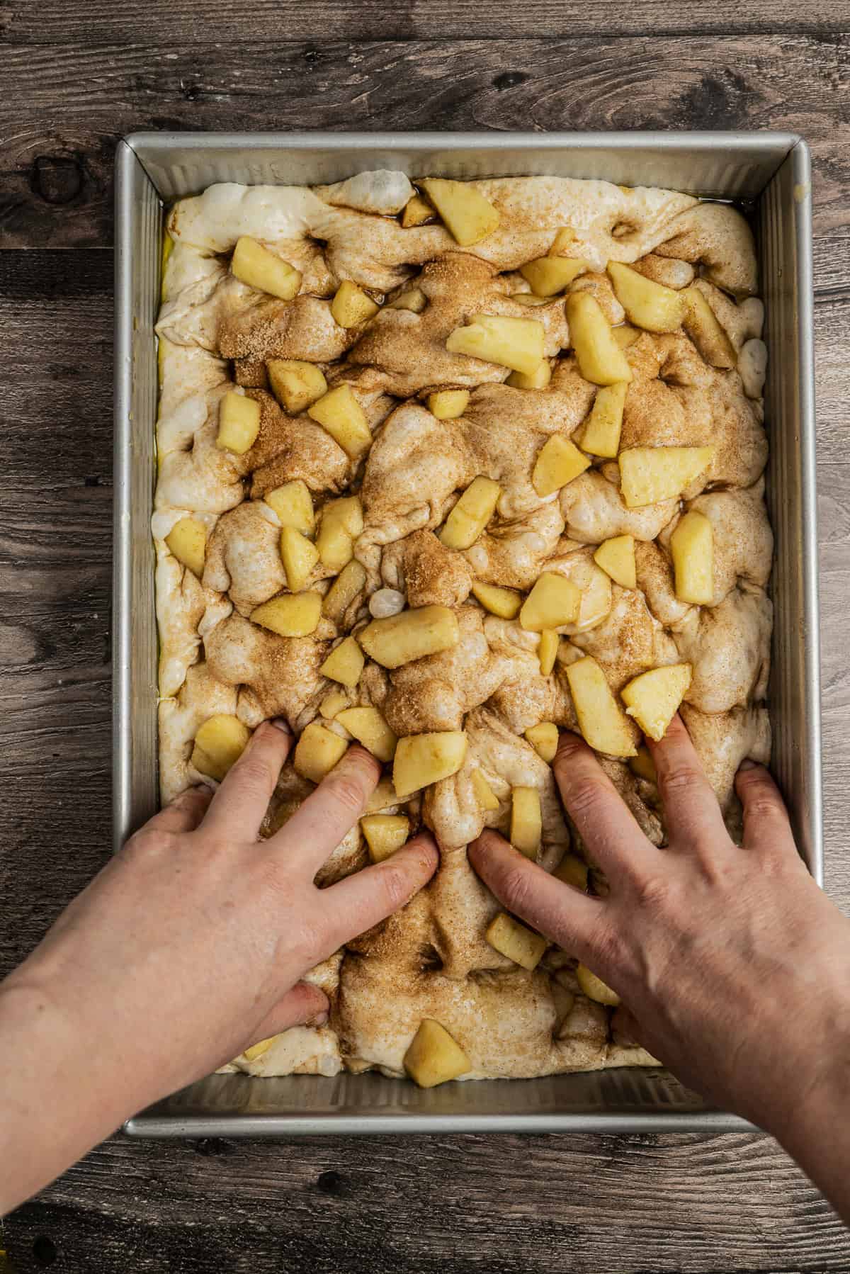 Hands pressing dough topped with cinnamon and apple chunks into a rectangular baking pan on a wooden surface.