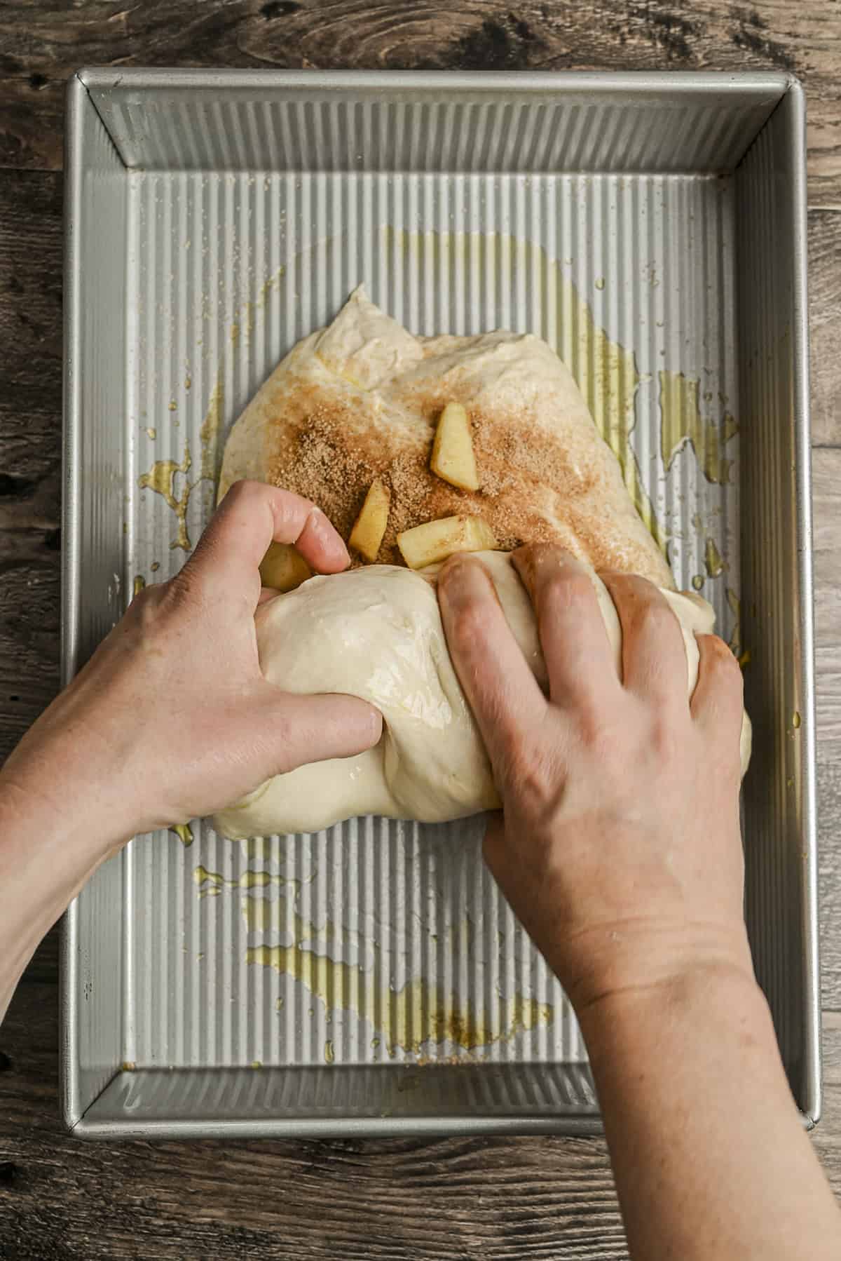 Hands folding dough with apple slices and cinnamon inside a greased metal baking pan on a wooden surface. The scene suggests preparation for baking a sweet bread or pastry.
