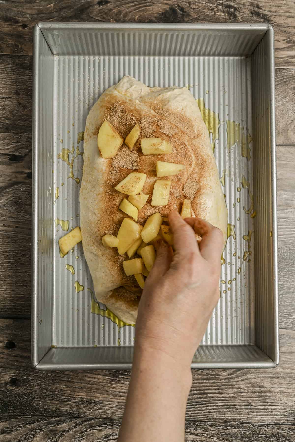 A hand sprinkles diced apples onto a sheet of pastry dough topped with cinnamon sugar, placed in a rectangular metal baking pan on a wooden surface.