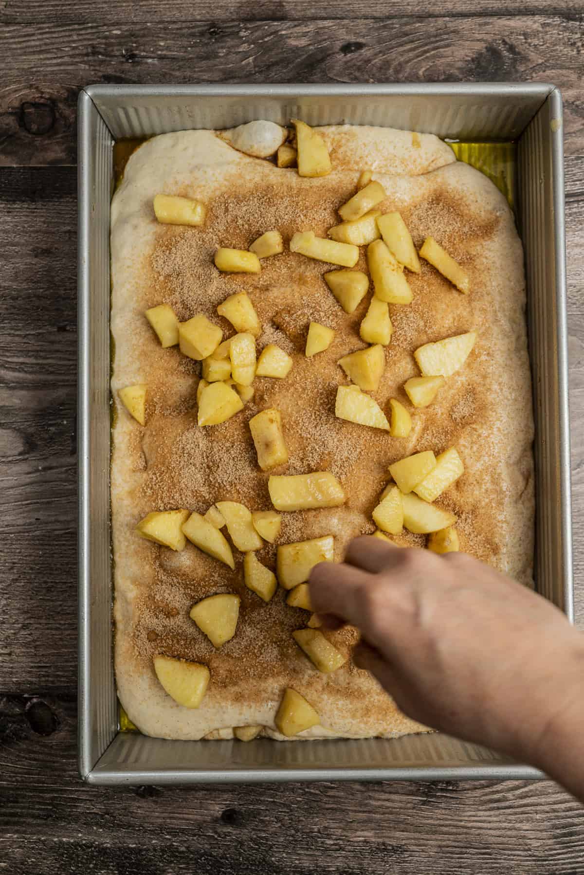 A hand sprinkles chopped apples over cinnamon-sugar covered dough in a rectangular baking pan on a wooden surface.