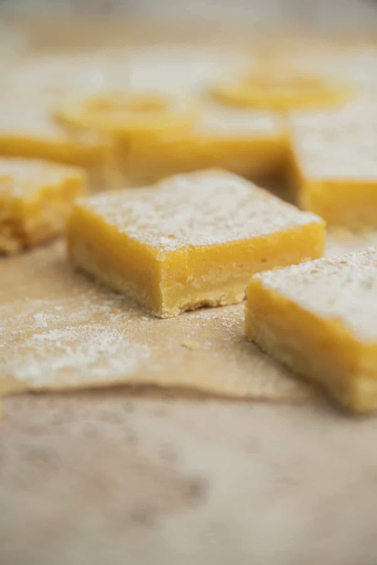 Close-up of lemon bars with a dusting of powdered sugar on top, arranged on a light-colored parchment paper surface. The bars have a golden yellow filling and a light crust.
