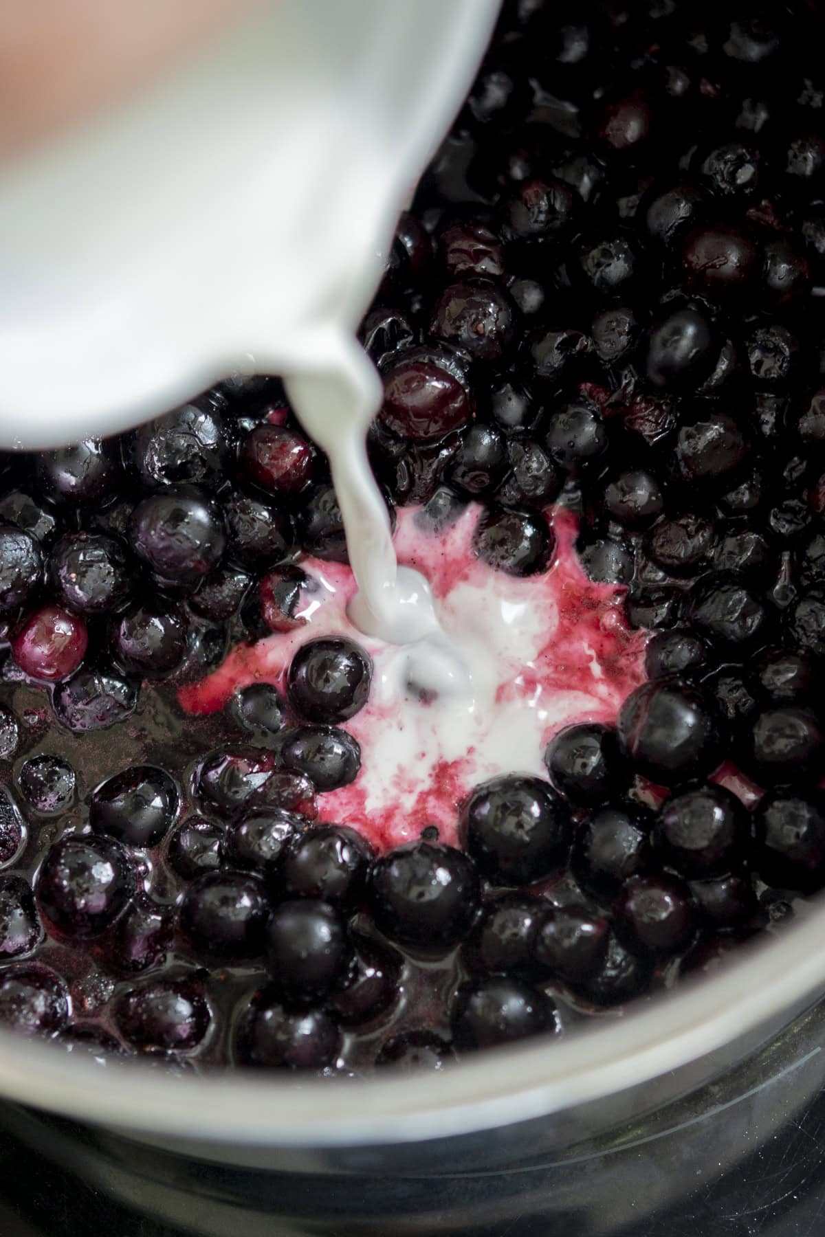 Close-up of a creamy white liquid being poured onto a pot of simmering blueberries, creating a pink swirl in the mixture.