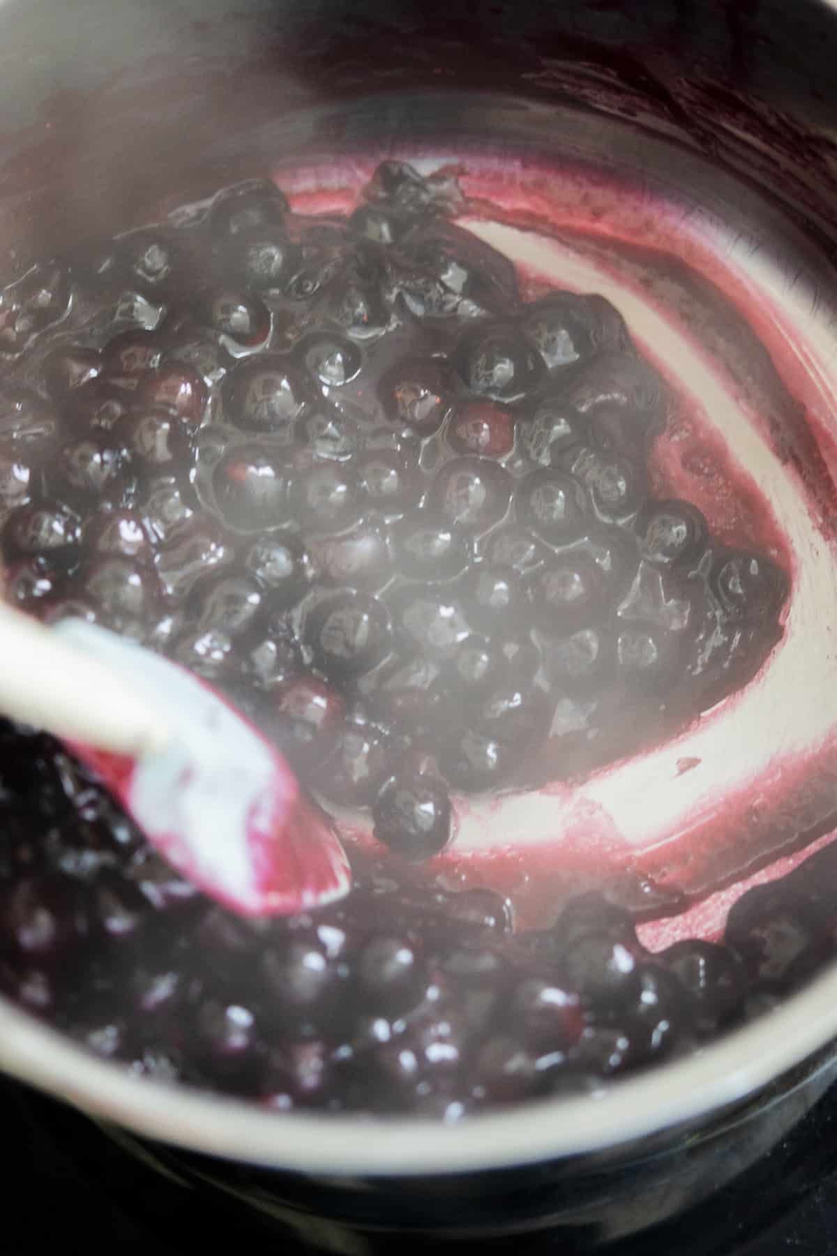 Blueberries are being cooked and stirred with a white spoon in a pot, creating a thick, purple mixture with visible steam rising.