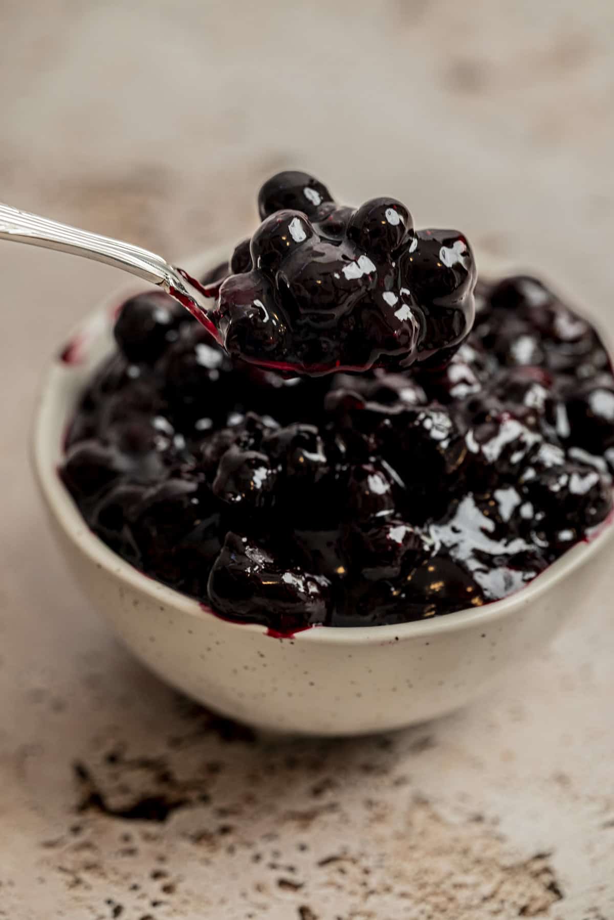 A close-up of a spoon lifting glossy, dark blueberries in syrup from a small, speckled bowl, set on a textured surface.