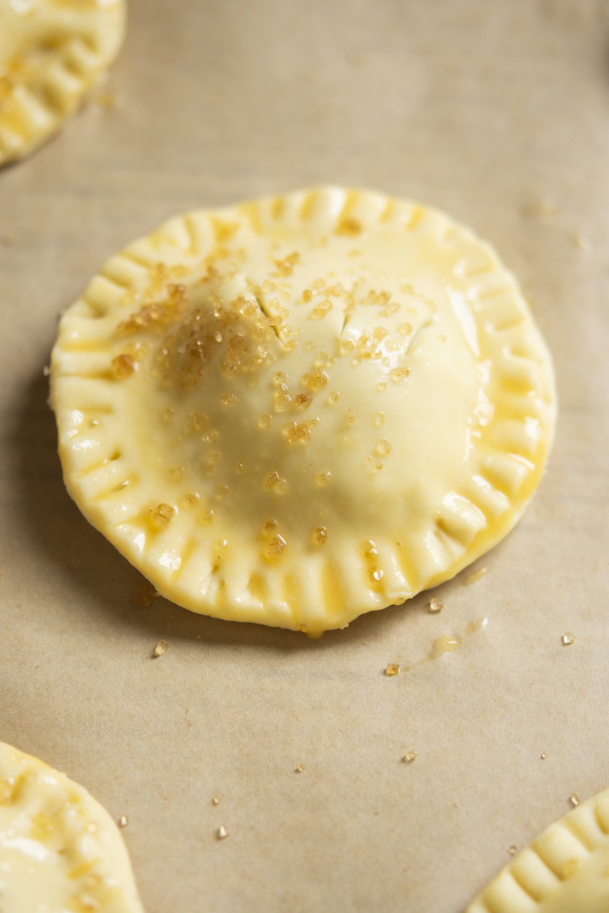 A close-up of an unbaked, round pastry with crimped edges, brushed with egg wash and sprinkled with coarse sugar, resting on parchment paper.