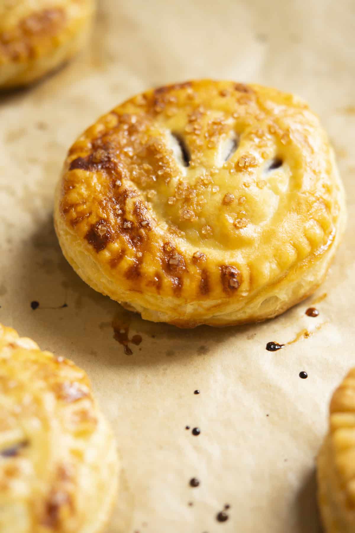 A golden-brown, round pastry with a smiling face design on top, resting on a sheet of parchment paper. Other similar pastries are partially visible in the background.