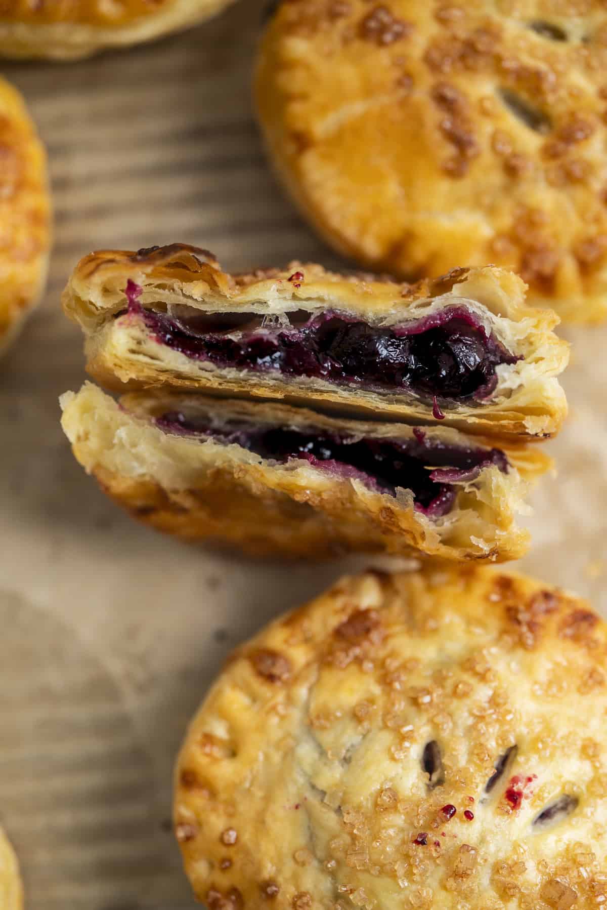 A close-up of golden, flaky hand pies, one of which is cut in half to reveal a blueberry filling. The pastries are sprinkled with coarse sugar and rest on a brown parchment surface.