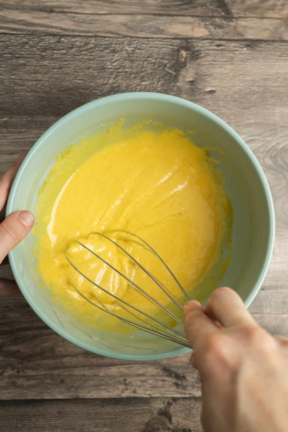 A person whisking yellow batter in a light blue bowl on a wooden surface.