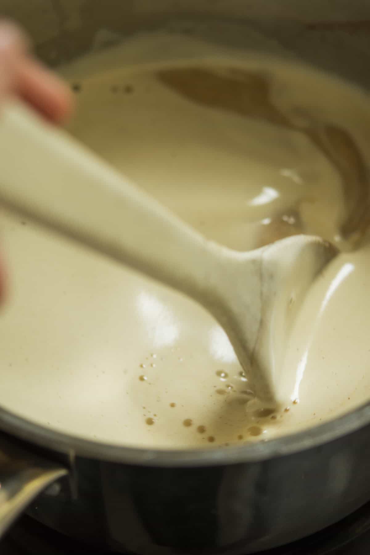 A close-up of a creamy beige sauce being stirred in a pan with a wooden spoon, creating smooth swirls and bubbles on the surface.