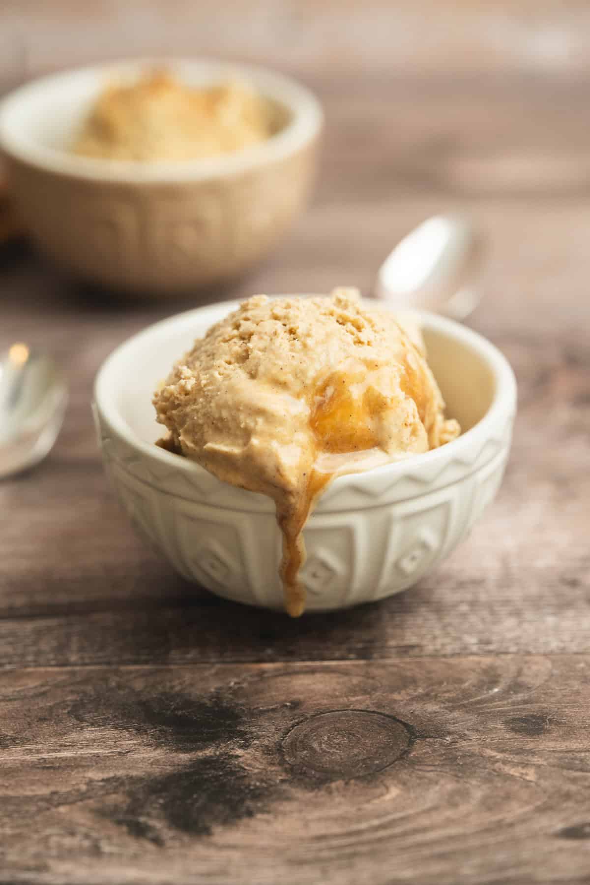 A scoop of creamy ice cream with caramel swirls in a patterned white bowl sits on a wooden surface, with another bowl and spoon blurred in the background.