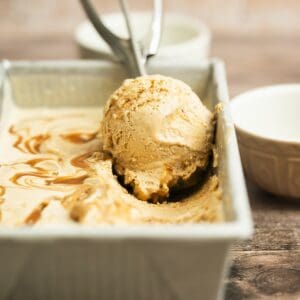 A close-up of a metal container with creamy brown caramel cinnamon ice cream, and a scoop resting in an ice cream scooper. A white bowl sits in the background on a wooden surface.