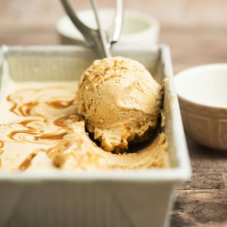 A close-up of a metal container with creamy brown caramel cinnamon ice cream, and a scoop resting in an ice cream scooper. A white bowl sits in the background on a wooden surface.