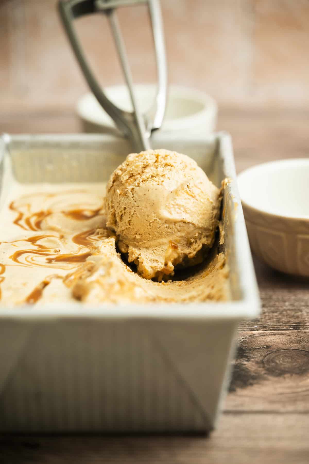 A metal container filled with creamy caramel-colored cinnamon caramel Cream, with a scoop resting on an ice cream scooper. Swirls of caramel sauce are visible on top. The scene is set on a wooden surface with bowls nearby.