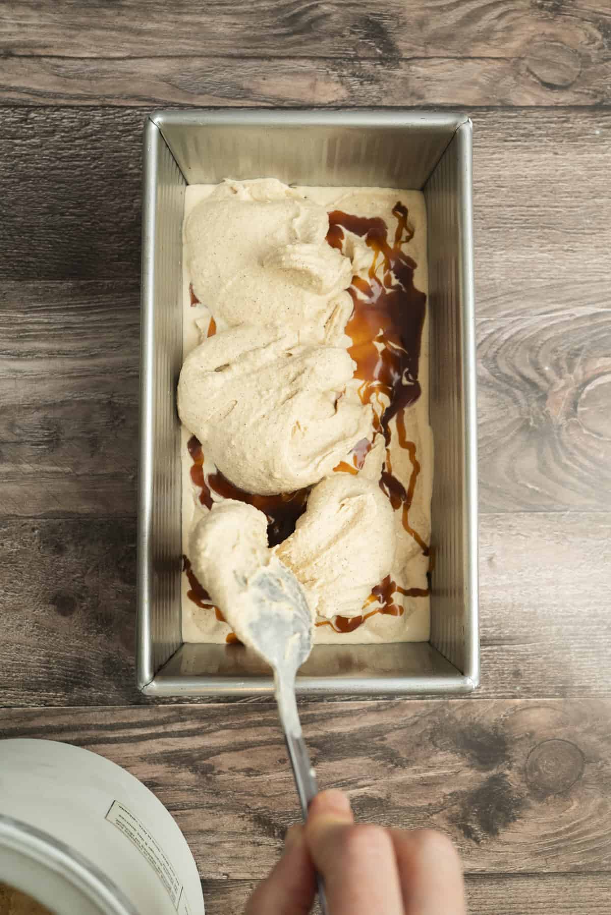 A hand uses a spoon to spread creamy ice cream mixture and caramel sauce in a rectangular metal loaf pan on a wooden surface.