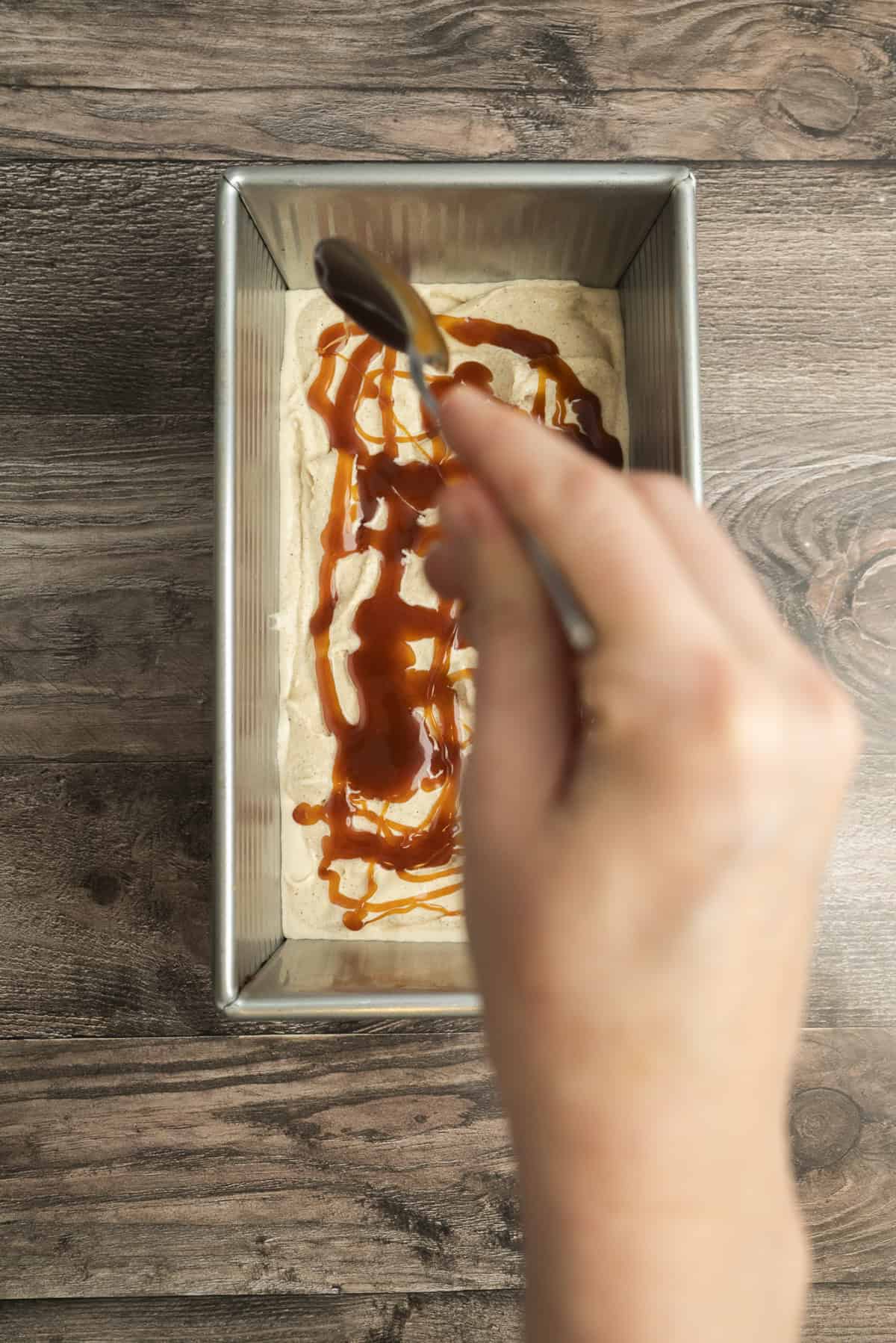A hand holding a spoon drizzles caramel sauce over a layer of ice cream mixture in a rectangular metal loaf pan, set on a wooden surface.