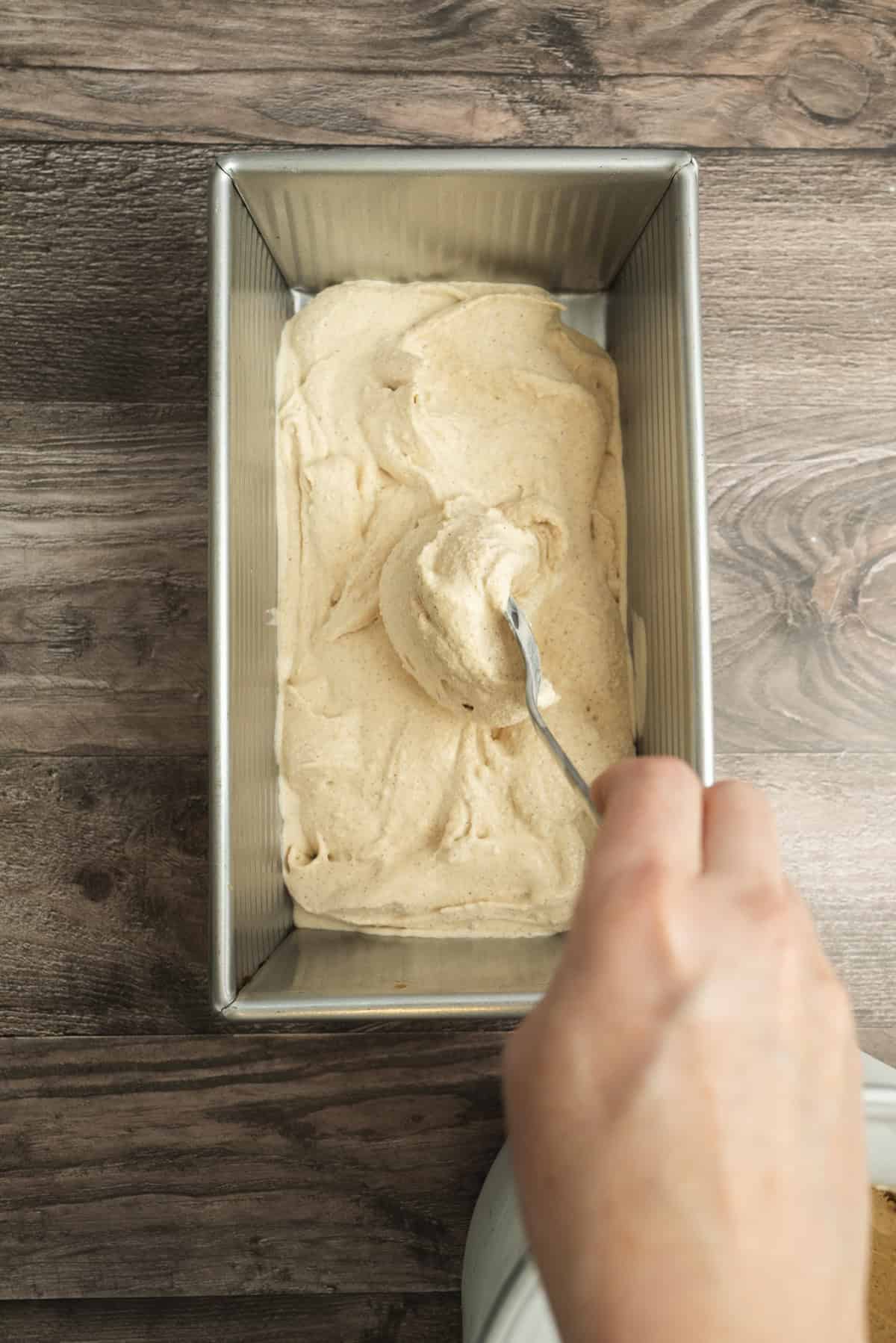 A hand spreads light brown ice cream or batter evenly in a metal loaf pan with a spoon, on a wooden surface.