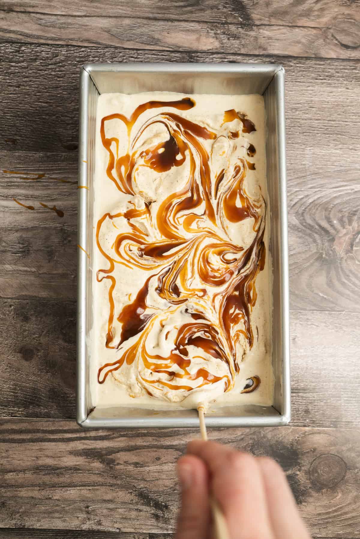 A hand uses a stick to swirl caramel into a creamy mixture in a rectangular metal pan, creating a marbled pattern. The pan sits on a wooden surface.