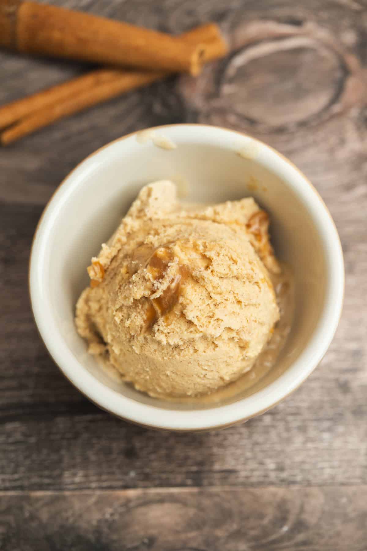 A scoop of light brown ice cream in a white bowl, topped with a drizzle of syrup, sits on a wooden surface with two cinnamon sticks in the background.