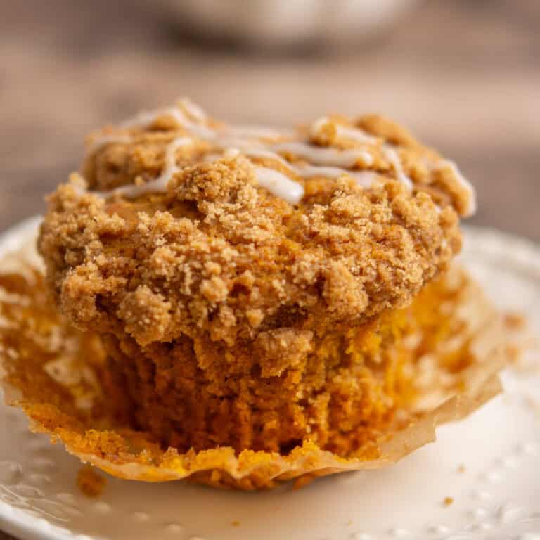 A close-up of a pumpkin muffin with a crumbly streusel topping and a drizzle of icing, partially unwrapped and sitting on a white plate.