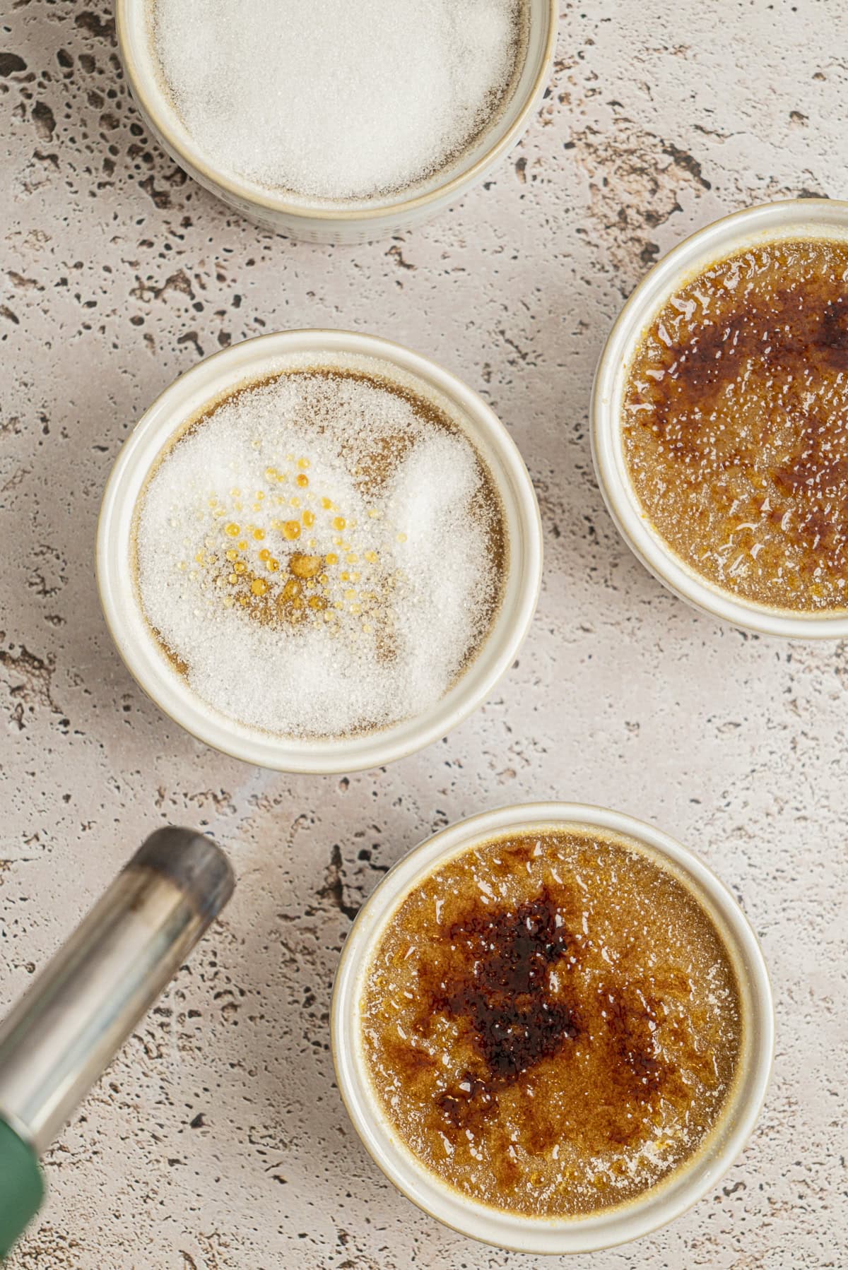 Four ramekins of crème brûlée sit on a textured surface; two have caramelized tops, one is being torched, and one is topped with granulated sugar, with a blowtorch visible in the lower left corner.