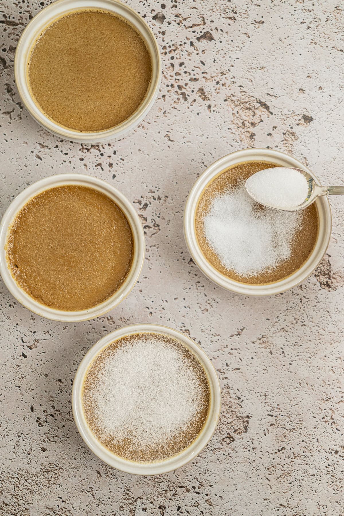 Four ramekins of crème brûlée are arranged on a textured surface. A spoon is sprinkling sugar onto one dessert, while another ramekin is already topped with a layer of sugar, ready to be caramelized.