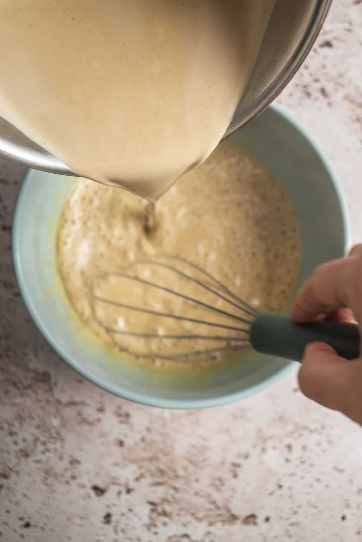 A hand holds a whisk in a blue bowl while a creamy liquid is being poured in from a metal bowl above, mixing ingredients on a light countertop.