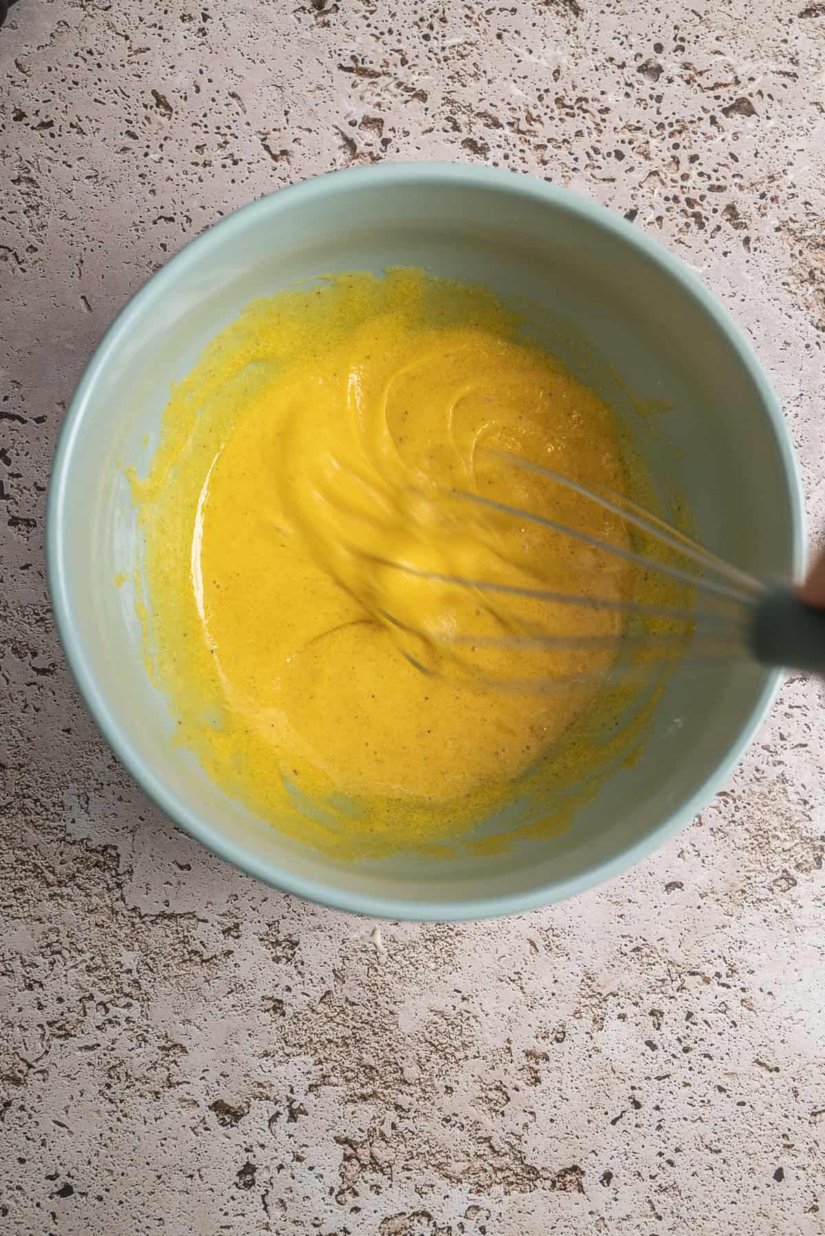 A hand whisks a egg yolks in a light blue bowl, placed on a textured, beige countertop.